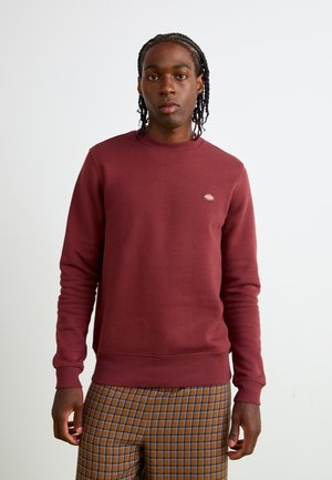 Bordeauxrode sweatshirt met geribde manchetten en zoom, voorzien van een klein merklong op de borst. Gecombineerd met bruine geruite broek.