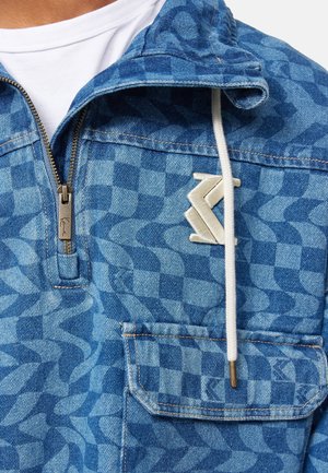 Personne en veste zippée bleue à motifs et pantalon assorti avec deux poches avant et cordons blancs, debout les mains jointes.