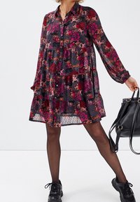 Robe à motifs floraux avec fond sombre, fermeture à boutons sur le devant, manches longues et design à volants ; portée avec des chaussures noires et un sac à main.