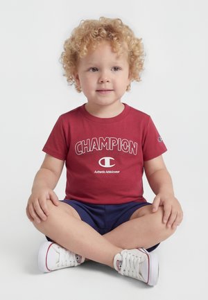 Ungt barn med krøllete hår som sitter med korslagte ben, iført en rød Champion t-skjorte, marineblå shorts og hvite joggesko på en ensfarget bakgrunn.