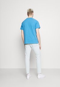 Persona con el cabello corto y decolorado, vistiendo una camiseta azul, jeans claros, calcetines blancos y zapatillas blancas, de pie frente a una pared lisa y clara.