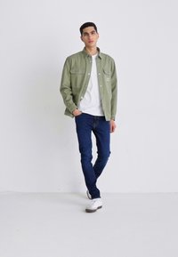 Veste verte à boutons sur un t-shirt blanc, associée à un jean slim bleu foncé et des baskets blanches, sur un fond uni.