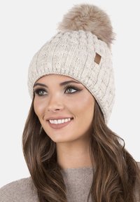 Gorro de punto beige con un patrón texturizado y un pompón esponjoso en la parte superior. Presenta una pequeña etiqueta de cuero en el lateral.