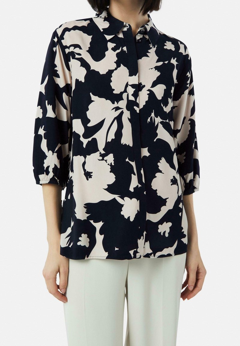 Femme portant un chemisier à boutons noir et blanc à motif floral avec des manches trois-quarts et un pantalon de couleur claire sur un fond uni.