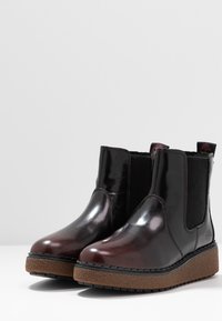 Brązowe, lakierowane botki chelsea z elastycznymi panelami bocznymi i teksturowanymi, brązowymi podeszwami. Zaokrąglony nosek i subtelne logo po boku.