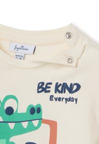 Cream cotton t-shirt with green crocodile graphic. Features the text "BE KIND Everyday" in navy, and snap closures at the shoulder.