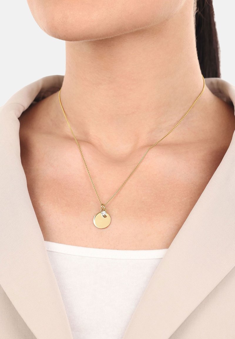 Collar de cadena de oro con un pequeño colgante redondo de oro y una única piedra preciosa clara, usado en el cuello de una mujer sobre una blusa blanca y un blazer beige.