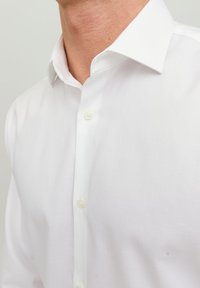 Hombre que lleva una camisa blanca de vestir impecable con cuello puntiagudo, abotonada hasta el cuello, sobre un fondo claro y liso.