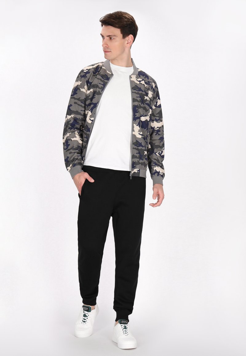 Giubbotto bomber in camouflage grigio, blu navy e crema, con zip, colletto a coste e pantaloni neri affusolati, abbinati a sneaker bianche.
