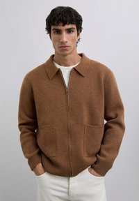 Mand med mørkt hår iført en brun lynlås-cardigan med krave og forlommer, hvid skjorte og hvide bukser, stående med hænderne i lommerne.