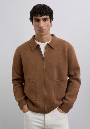 Hombre con cabello oscuro vistiendo un cárdigan marrón con cremallera y cuello, con bolsillos frontales, camisa blanca y pantalones blancos, de pie con las manos en los bolsillos.