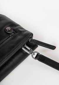 Schwarze Ledertasche mit einer glatten Textur, ausgestattet mit einem Reißverschluss, metallischen Akzenten und einem abnehmbaren Tragegurt mit Markenlogo.