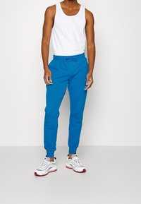 Blue jogger pants with an elastic waistband and drawstring, paired with a white tank top and white sneakers with red accents.