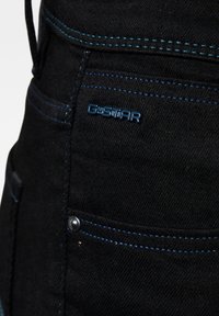 Material de mezclilla negra con costuras azules contrastantes. Presenta un logo de G-Star y un pequeño bolsillo con un botón de metal. Vista en primer plano.