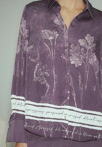 Lilla skjorte med knapper og blomsterprint samt hvid tekst ved kanten. Fremstillet af glat stof med krave og lange ærmer.