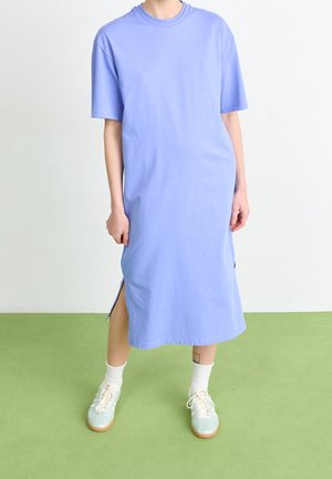Osoba dorosła ubrana w jasnoniebieską midi sukienkę typu t-shirt z krótkimi rękawami i rozcięciami po bokach, białe skarpetki i jasnozielone sneakersy na sznurowadła.