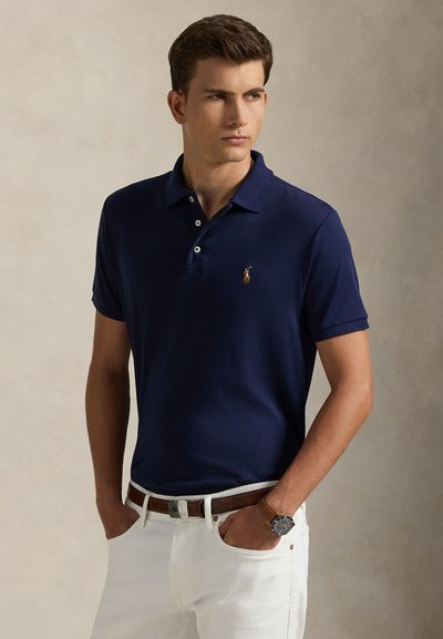 Junger Mann trägt ein dunkelblaues Poloshirt, weiße Hose, braunen Gürtel und Armbanduhr, steht mit den Händen in den Taschen und schaut zur Seite.