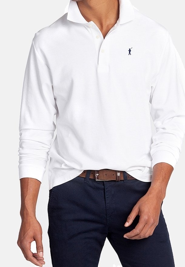 Homme portant un polo blanc à manches longues avec un petit logo bleu marine sur la poitrine gauche, associé à un pantalon foncé et une ceinture marron.