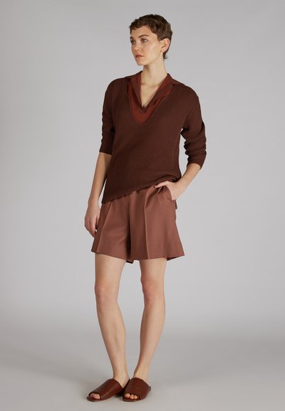 Maglione marrone a costine con scollatura a V profonda, indossato sopra una camicia a chevron rossa. Accompagnato da pantaloni corti marroni abbinati e sandali slip-on.