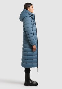 Padded long coat in light blue, featuring a hood, front zipper, and elastic cuffs. Worn with black ankle boots.