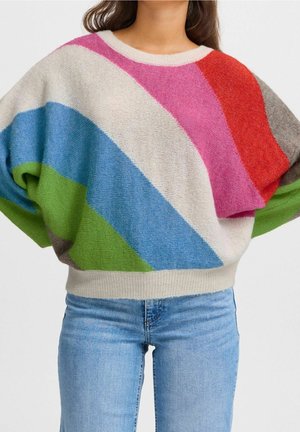 Femme portant un pull ample multicolore à rayures diagonales crème, rose, rouge, bleu, vert et gris, associé à un jean bleu clair.