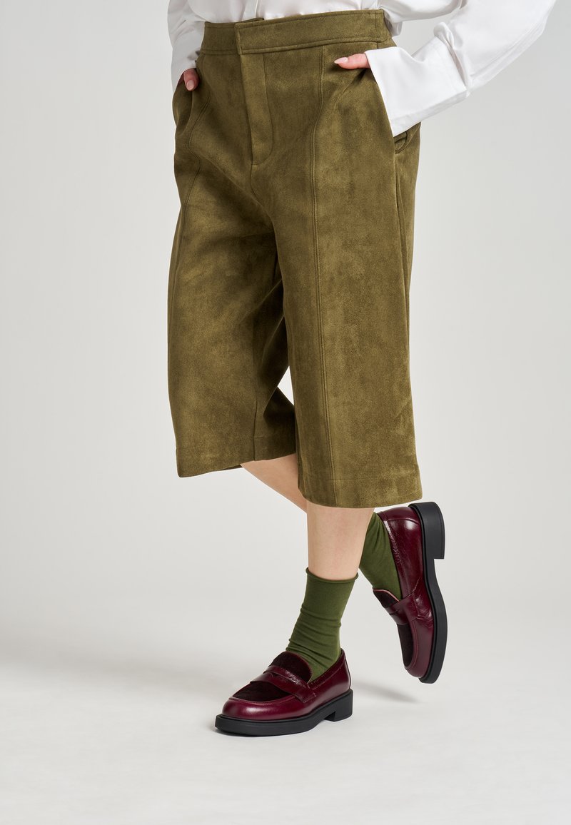 Culotte in camoscio verde oliva con parte anteriore piatta, tasche laterali e dettagli cuciti. Abbinati a mocassini bordeaux e calze verdi.