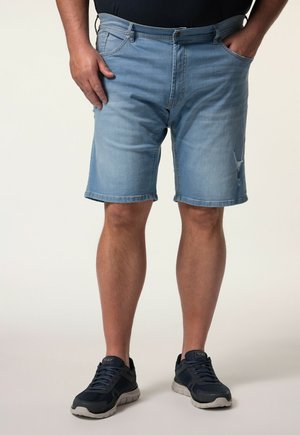 Mann trägt hellblaue Jeansshorts und schwarze Turnschuhe und steht mit den Händen in den Taschen vor einem schlichten Hintergrund.