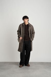 Brauner, strukturierter langer Mantel über einem dunklen Pullover, kombiniert mit schwarzen Hosen und braunen Schuhen, während er eine hellgraue Schultertasche hält.