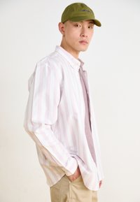 Camisa de botones a rayas en rosa claro y blanco sobre una camiseta color malva, combinada con pantalones caqui y una gorra verde oliva, con un ajuste casual.