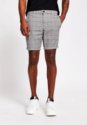 Mann trägt schwarz-weiß karierte, hochgekrempelte Shorts, schwarzes Hemd und weiße Sneakers, steht mit Händen in den Taschen vor weißem Hintergrund.