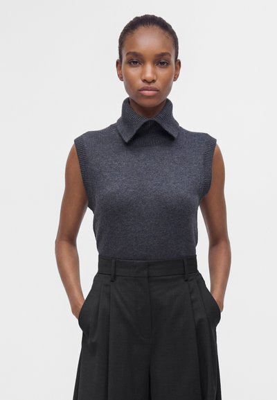 Femme à la peau foncée portant un haut en tricot sans manches gris foncé avec un col montant et un pantalon noir taille haute à jambes larges, les mains dans les poches.