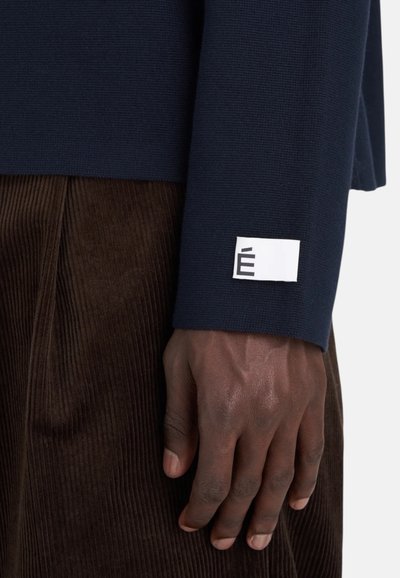 Haut à manches longues côtelé marine avec une étiquette blanche arborant un "É" stylisé sur le poignet, associé à un tissu en velours côtelé brun.