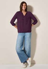 Blouse violette à coupe ample avec un col en V et un cordon de serrage. Manches trois-quarts et ourlet froncé. Associée à un jean en denim bleu clair à jambes larges et des baskets blanches.