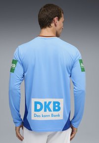 Helle blaue Sportjacke mit langen Ärmeln und dunkelblauen Akzenten. Enthält Sponsorenlogos in grün und einen großen weißen Aufdruck auf dem Rücken.