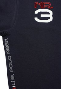 Granatowa koszulka z okrągłym dekoltem, z czerwonymi i białymi grafikami: "NR. 3" nadrukowane centralnie oraz "U.S. POLO ASSN." wzdłuż boku na biało.