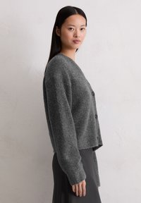 Grijze, gestructureerde cardigan met een ronde halslijn, lange mouwen en knopen aan de voorkant. Draagt over een donkere, vloeiende rok.