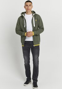 Green zip-up hoodie with white drawstrings and yellow accents, paired with a white T-shirt and dark jeans. Black sneakers complete the look.