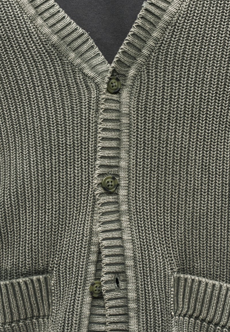 Nahaufnahme eines grauen Strickcardigans mit gerippten Kanten, drei dunklen Knöpfen und zwei vorderen Taschen über einem dunklen Unterhemd.