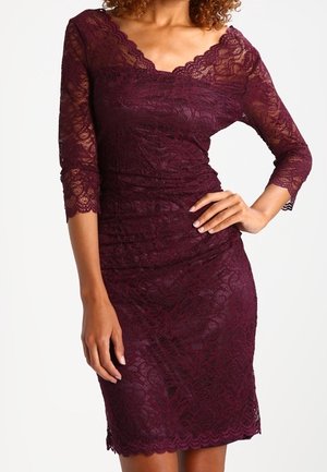 Kobieta ubrana w dopasowaną, bordową koronkową sukienkę do kolan z rękawami 3/4 i falbankowatymi krawędziami, pozująca z jedną ręką na biodrze.