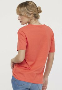 Korallefarbenes Baumwoll-T-Shirt mit rundem Ausschnitt und kurzen Ärmeln, von hinten gesehen, kombiniert mit blauen Jeans und lässigem Schnitt.