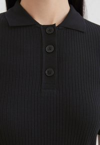Zwarte geribde poloshirt met een kraag en drie zwarte knoopsluitingen aan de voorkant, gestructureerde stof met verticale lijnen.