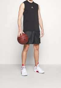 Maglietta nera senza maniche, pantaloni corti in rete neri con accenti bianchi e scarpe da basket bianco-rosse. Tiene in mano un pallone da basket con un logo.