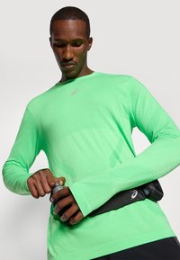 Grønn langermet treningstrøye med stretch-stoff, med rund hals og sømløst design. En svart midjeveske holder en flaske.