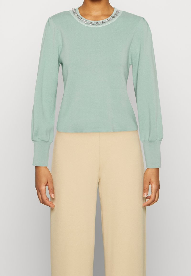 Minzgrüner Pullover mit langen Puffärmeln und einem perlenverzierten Ausschnitt. Glatte Textur, figurbetontes Design, kombiniert mit cremefarbenen Hosen mit weitem Bein.