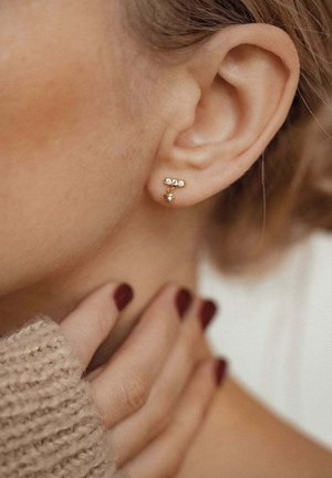 Goldohrringe mit drei klaren Edelsteinen, die in einer horizontalen Linie über einer herabhängenden goldenen Perle angeordnet sind. Tragen am Ohrläppchen.