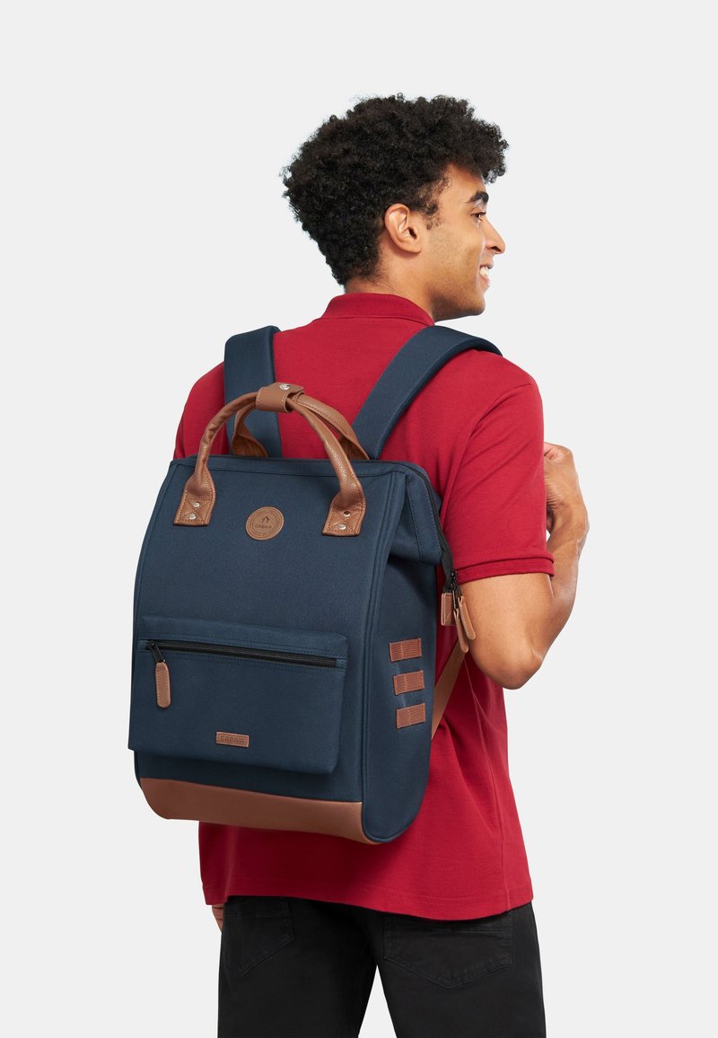 Donkerblauwe rugzak met bruine accenten, met een ritsvak aan de voorkant, een handvat bovenop en gevoerde verstelbare schouderbanden. De stof heeft een gladde textuur.
