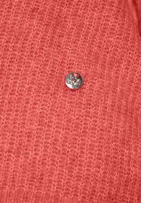 Texturerad ljusrosa tröjtyg med en silverfärgad cirkulär tagg med "MOS MOSH" präglad. Designen visar en mjuk, vävd look.