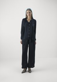 Blusa di seta blu navy con bottoni, abbinata a pantaloni blu navy larghi. Scarpe grigie a punta. Texture liscia, design minimalista.