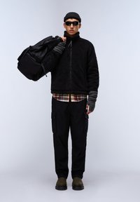 Giacca a zip nera in pile, camicia a quadri, pantaloni cargo neri, stivali verde oliva, guanti grigi senza dita, borsa a tracolla nera e occhiali da sole.