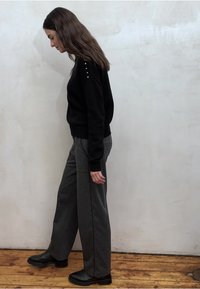 Een zwart gebreide trui met knoopdetails op de schouder, gecombineerd met grijze wijde broekspijpen en zwarte enkellaarzen op een houten vloer.